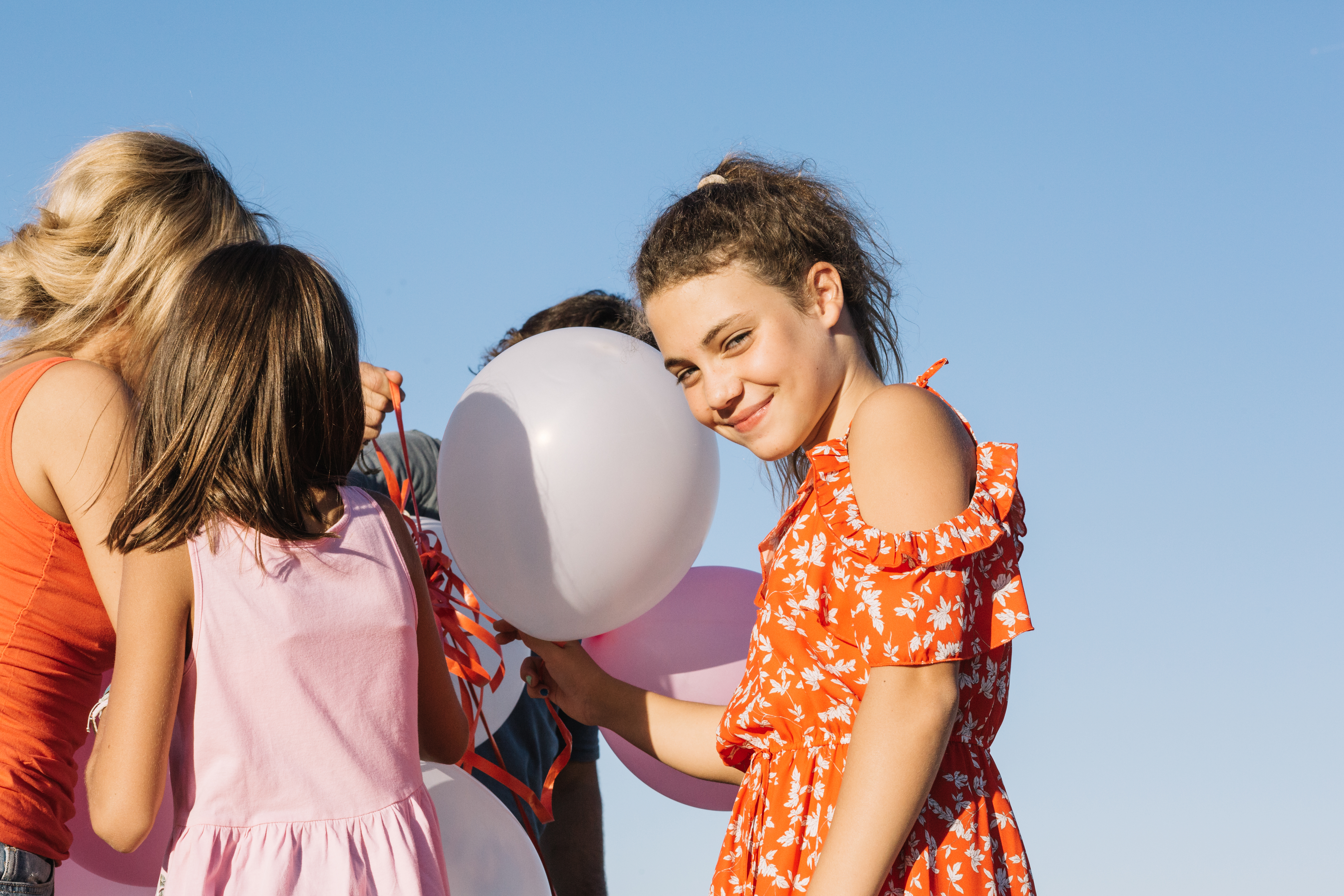 A smiling girl in an orange dress holding balloons against a blue sky